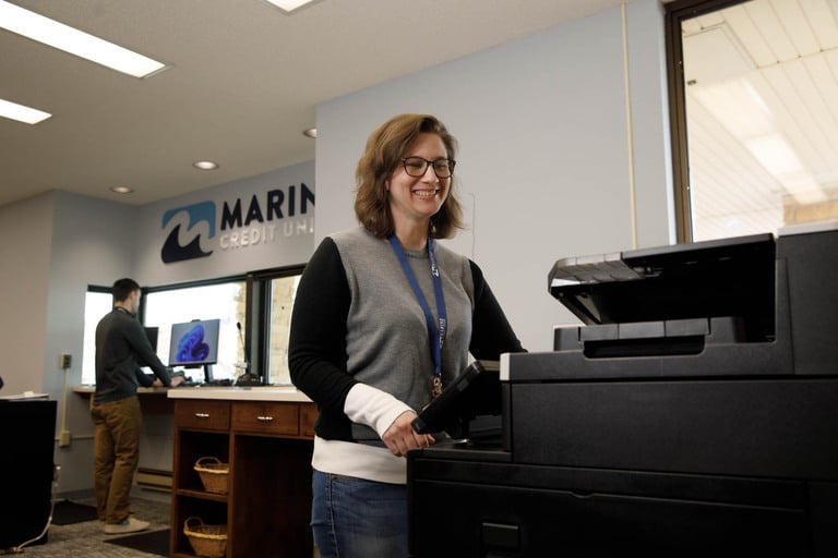 an image of a bank teller using a printer while another teller helps a customer in the background at marine credit union in chippewa falls wisconsin